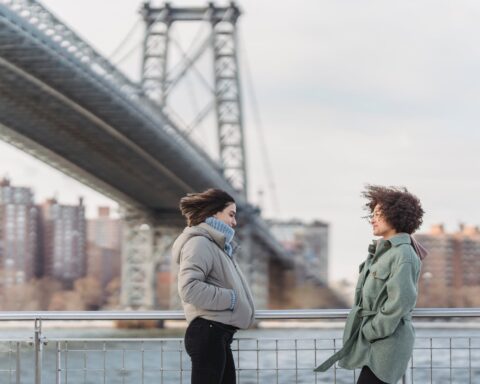 women in outerwear chatting on city embankment on windy day