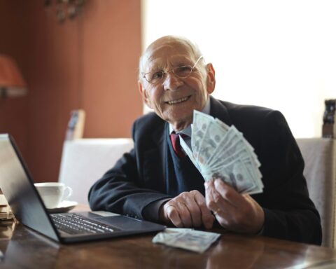 happy senior businessman holding money in hand while working on laptop at table