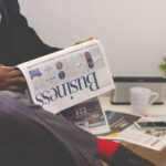 man reading newspaper while sitting near table with smartphone and cup